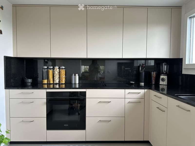 Modern kitchen with light-colored cabinets, a built-in oven, and a sleek black countertop. The countertop features pasta jars, a blender, and other kitchen items.