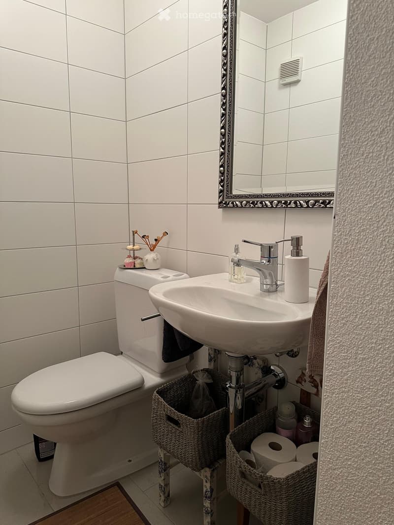 Modern bathroom with white tiles, a toilet, a sink with a chrome faucet, a mirror with a decorative frame, and wicker baskets holding toilet paper and other items.