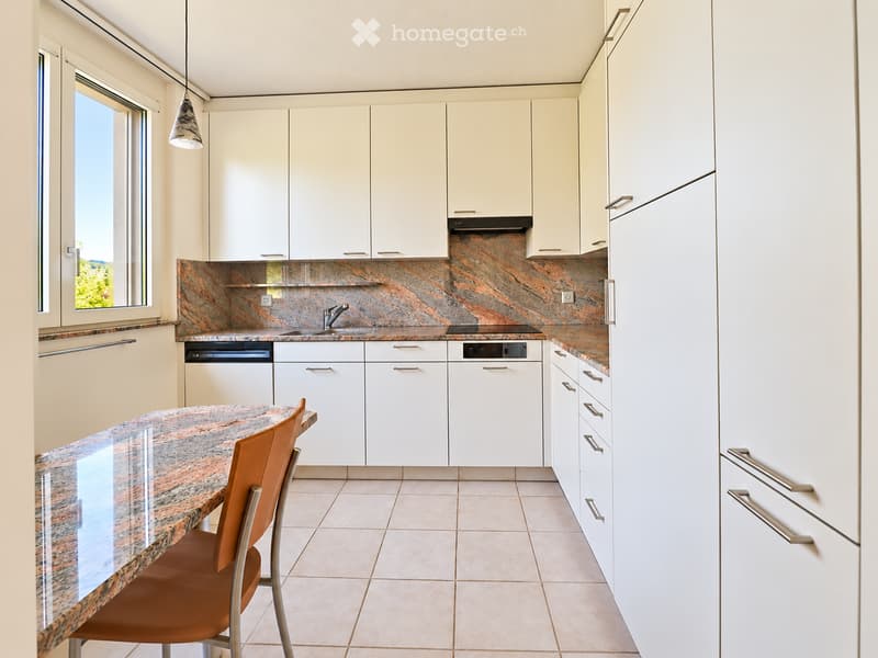 Modern kitchen with white cabinets, marble countertops, a central island, and a single chair. Features a sink, dishwasher, and stove.