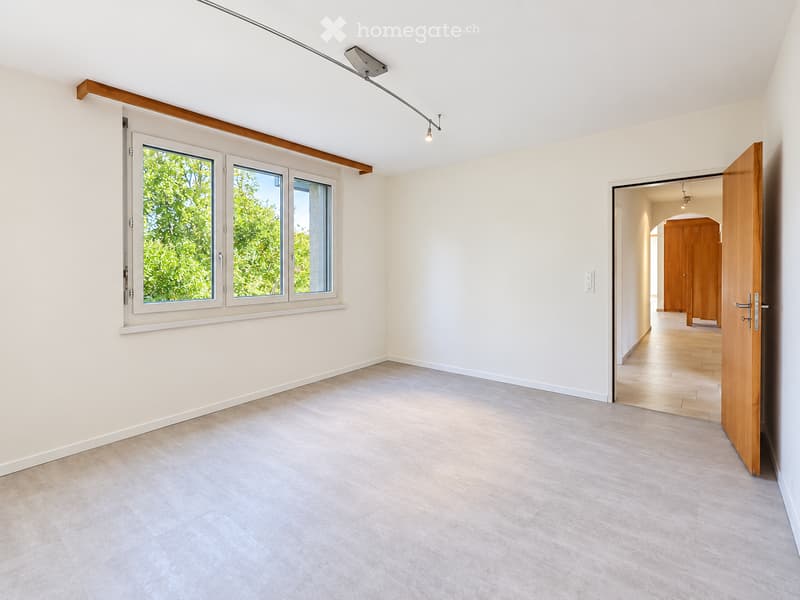 Empty room with light-colored walls, tiled floor, and large windows showing greenery outside. A wooden door leads to another room with wooden flooring and built-in cabinets.