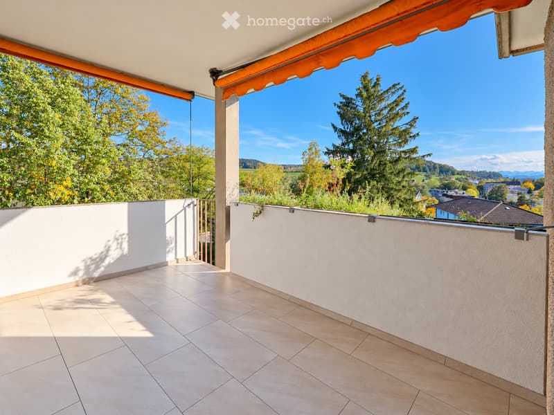 Balcony with tiled floor, white walls, metal railing, and an orange awning, overlooking a scenic landscape with trees and distant hills.