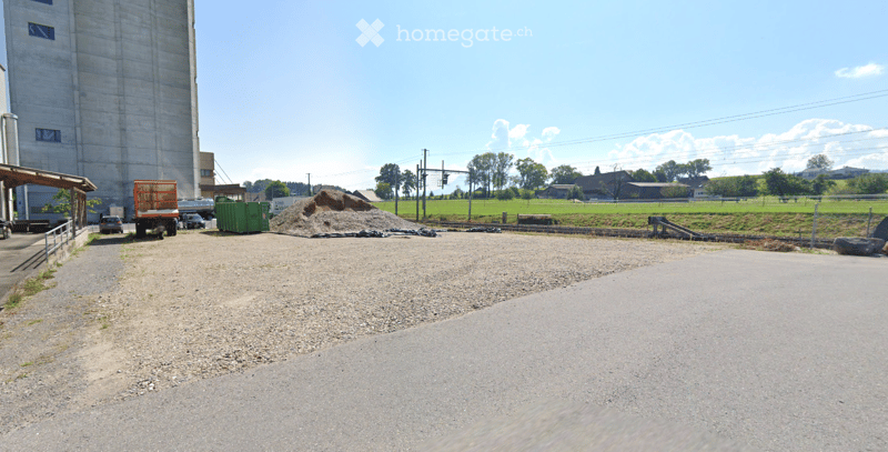 Gravel driveway leading to a construction area with a large pile of dirt, a truck, and a green container. A tall concrete silo is visible in the background, with a grassy field and scattered trees beyond.