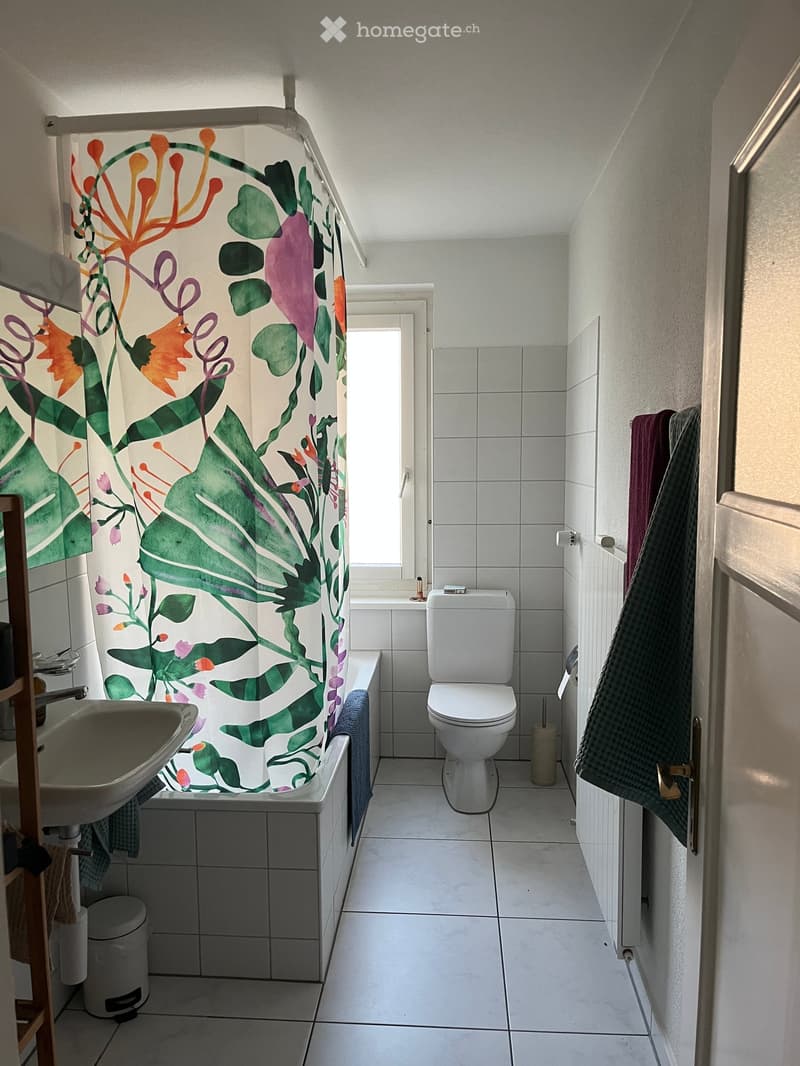 Bathroom with white tiles, a toilet, a bathtub with a colorful shower curtain, a sink, and towels hanging on the wall.