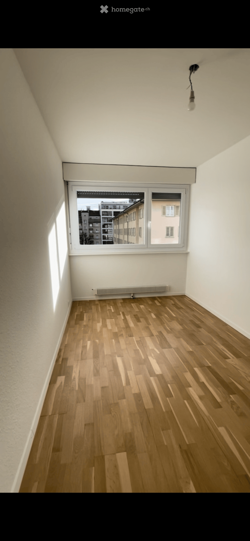 Empty room with wooden flooring, white walls, and a large window showing a cityscape view. No furniture or equipment present.