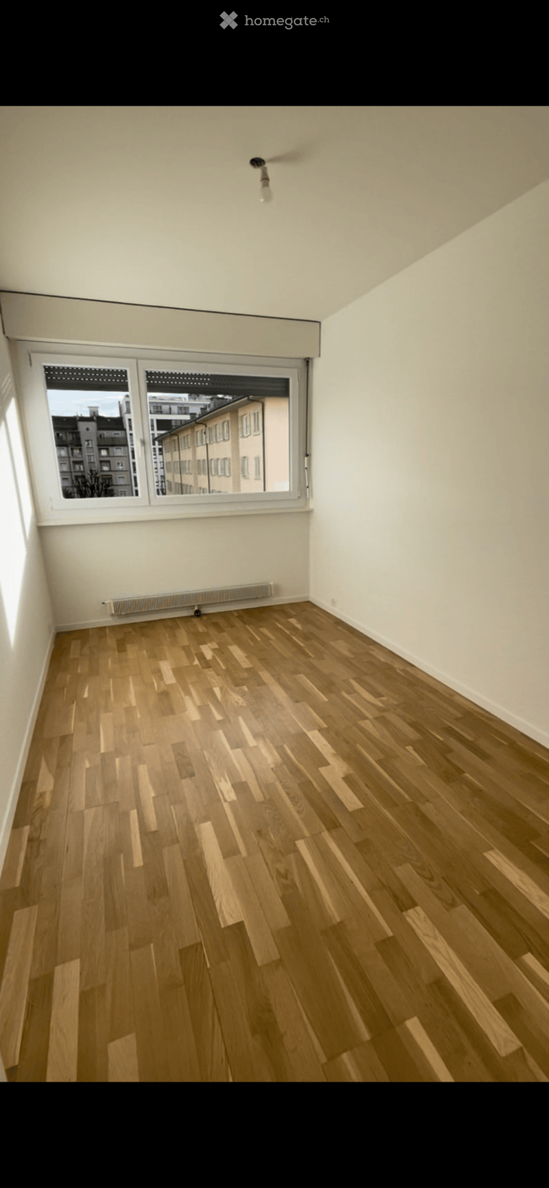 Empty room with wooden flooring, white walls, and large windows offering a view of the cityscape.