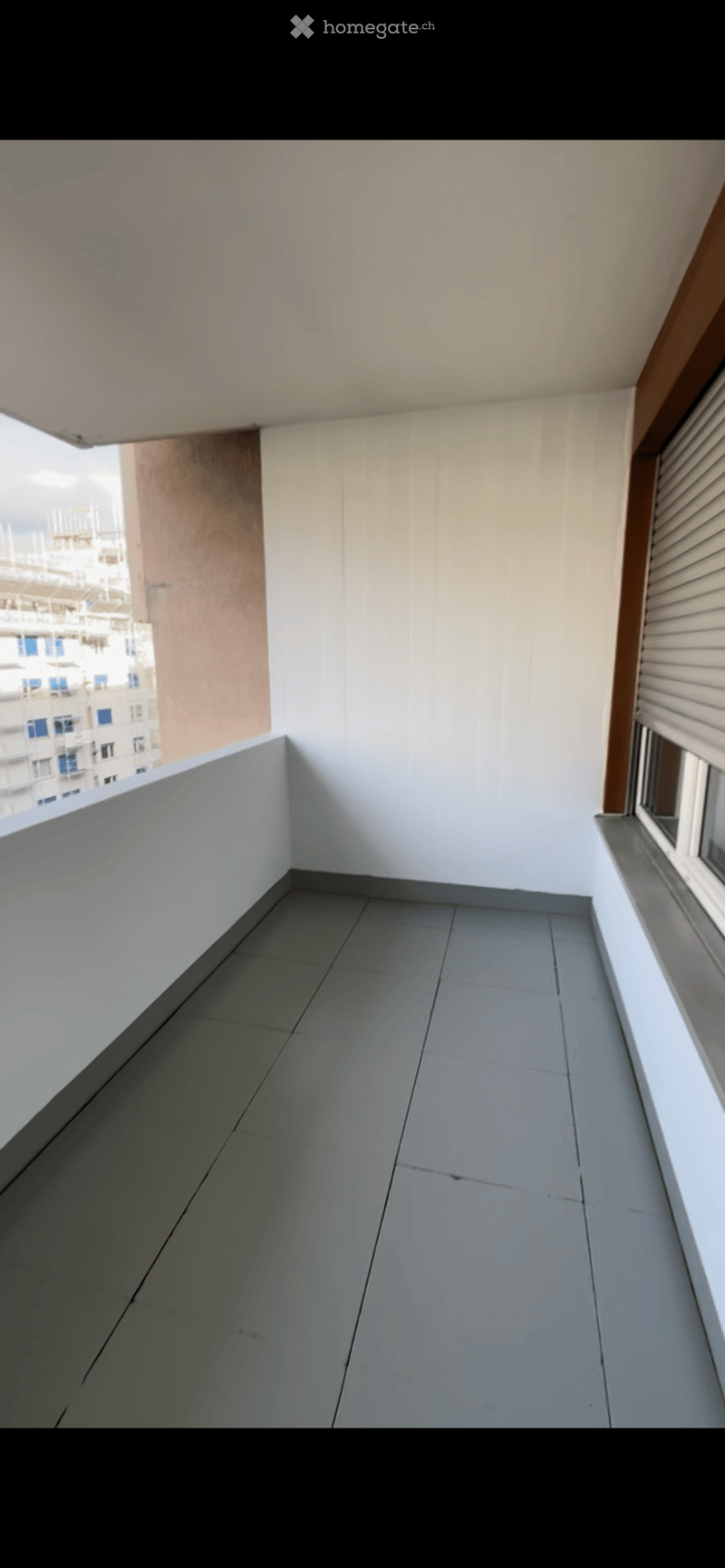 Empty balcony with grey tiled floor, white walls, and a view of neighboring buildings.