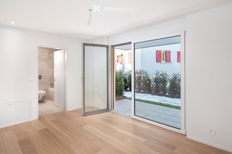 Empty room with light wooden flooring, white walls, and large glass sliding doors leading to an outdoor patio. A bathroom is visible through an open doorway.