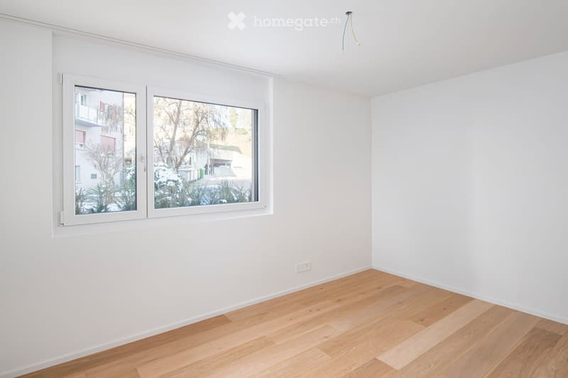 Empty room with white walls, wooden floor, and large windows showing a view of the street and neighboring buildings.