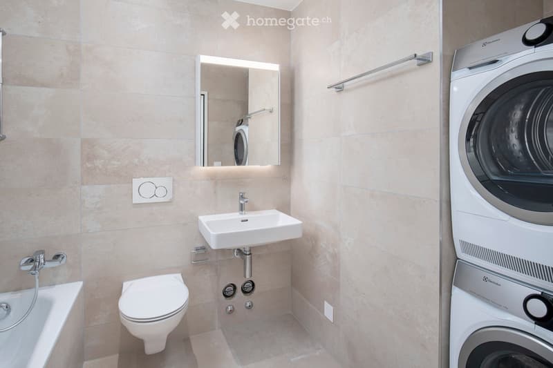 Modern bathroom with beige tiles, a white sink, toilet, bathtub, and a built-in washing machine. Features a large mirror with built-in lighting and a towel rack.