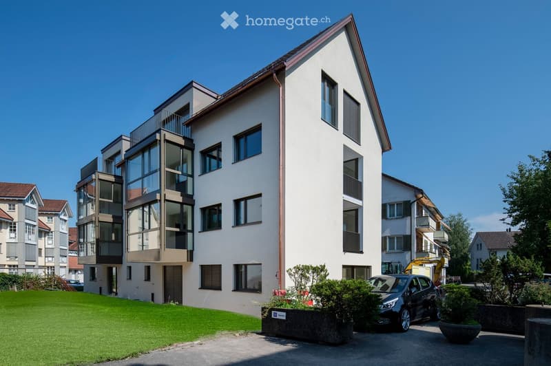 Modern multi-story residential building with white facade, large windows, and balconies. Features a well-maintained lawn, parked car, and surrounding greenery.