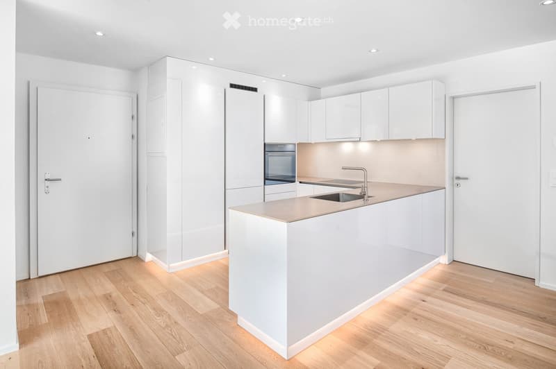 Modern, minimalist kitchen with white cabinets, a central island, and a sink with a faucet. The floor is light wood, and the walls are white.