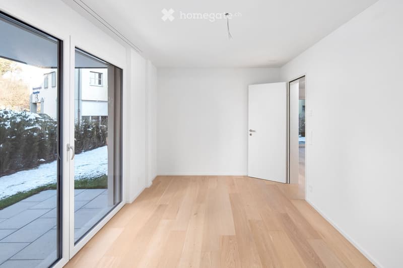 Empty room with light wooden flooring, white walls, large glass sliding doors leading to a patio, and an open doorway to another room.