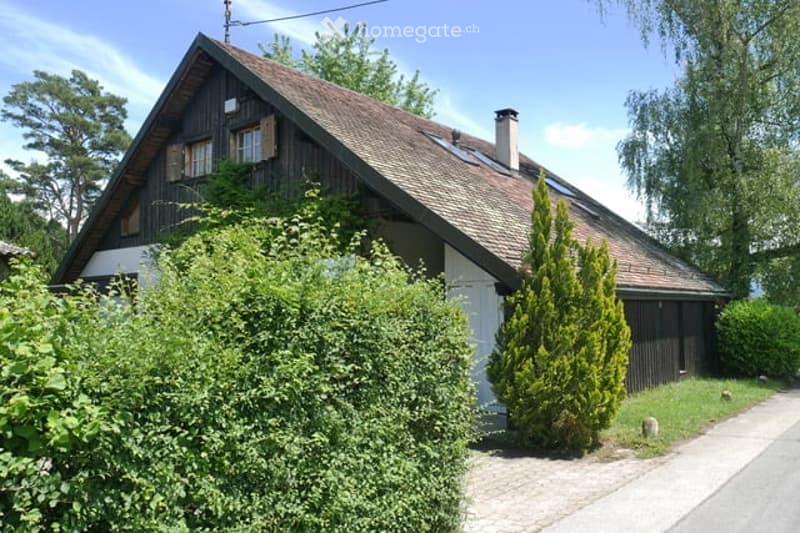 Traditional wooden house with a steeply pitched roof, surrounded by greenery and bushes.