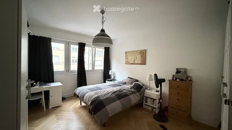 A well-lit bedroom with wooden flooring, a large window with dark curtains, a bed with a checkered duvet, a nightstand with a lamp, a fan, a desk with a chair, and a dresser with drawers.