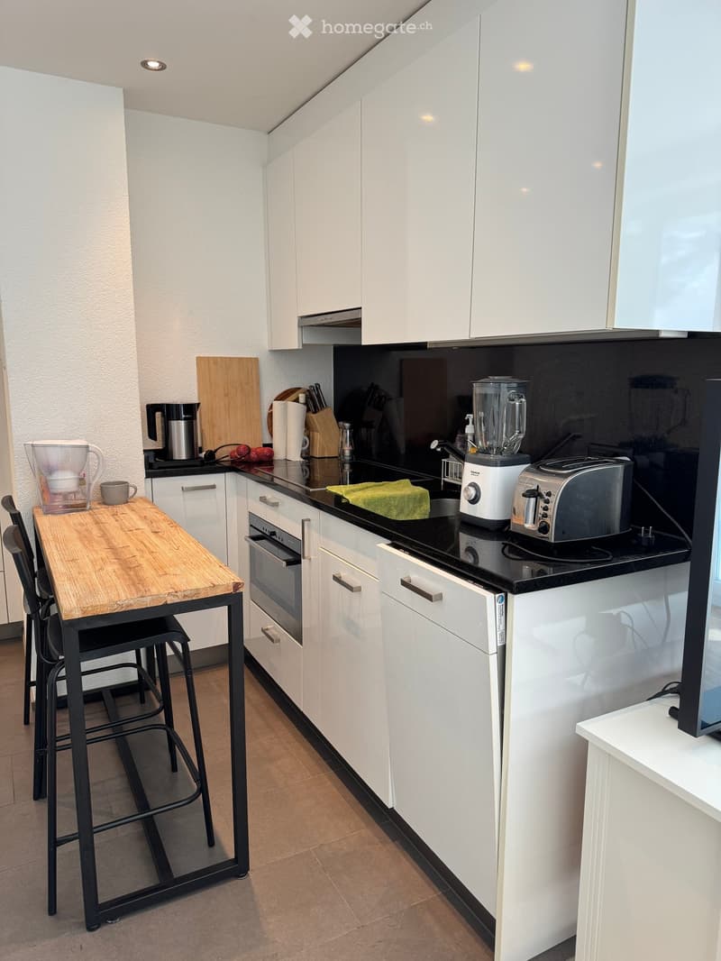 Modern kitchen with white cabinets, black countertop, wooden table, stools, and various appliances including a blender, toaster, and oven.