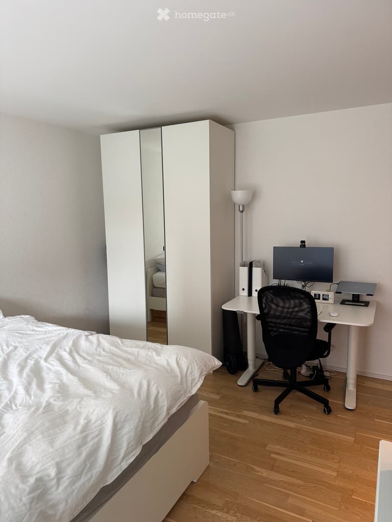 A modern, well-lit bedroom with a large bed, a white wardrobe with mirrored doors, a desk with a monitor, a chair, and a floor lamp.