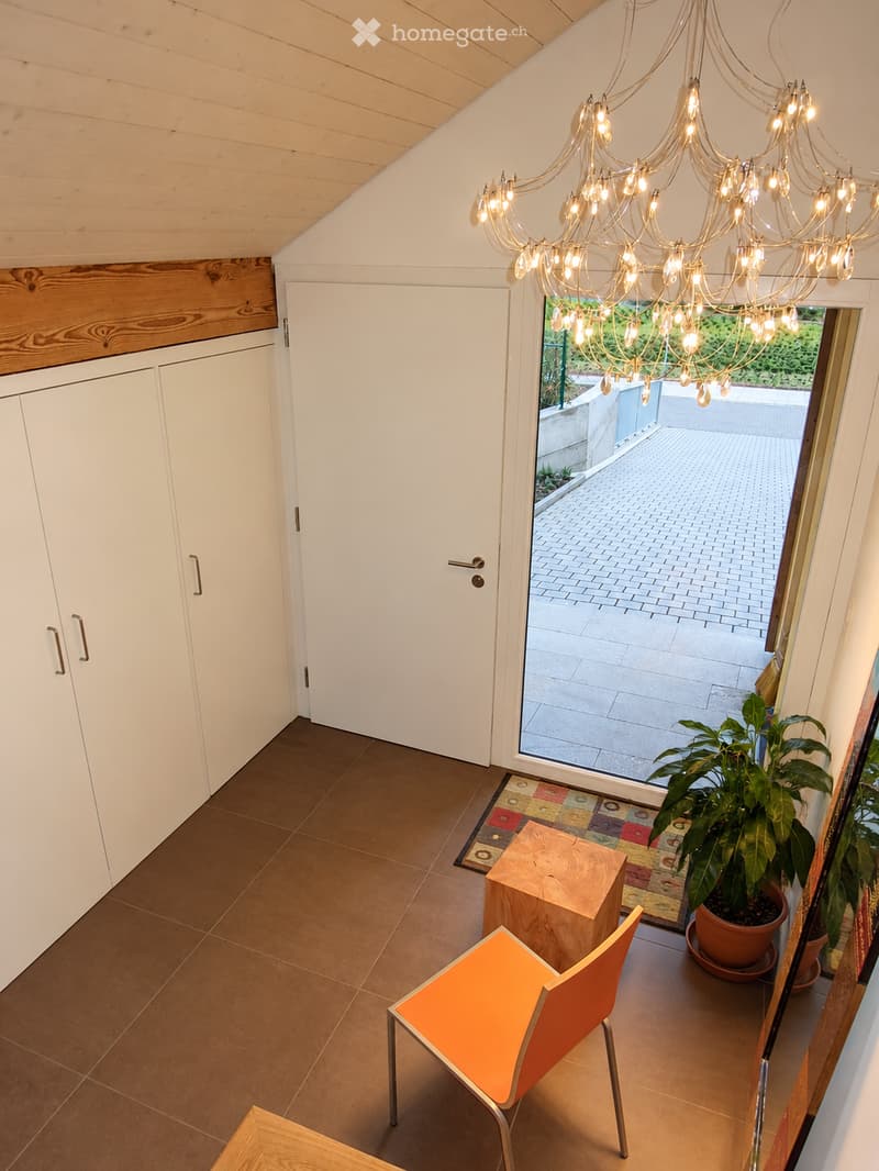 Bright, modern room with wooden ceiling beams, large glass door leading to patio, colorful rug, potted plant, and modern furniture including a bright orange chair and wooden stool.