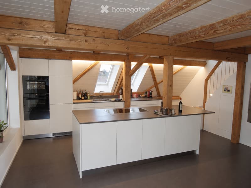 Modern kitchen with white cabinets, dark countertops, exposed wooden beams, and skylights. Features an island with a sink, oven, and various kitchen items on the counter.