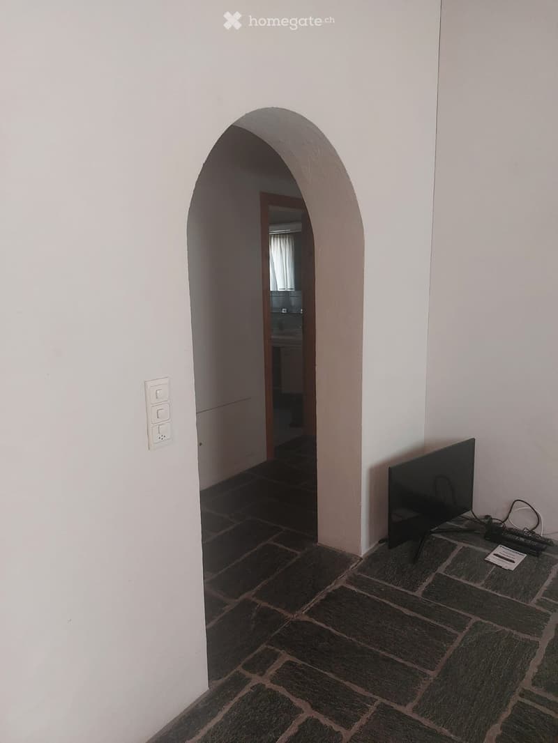 A room with a stone-tiled floor, white walls, and an arched doorway leading to another room. A flat-screen TV is placed on the right side of the room.