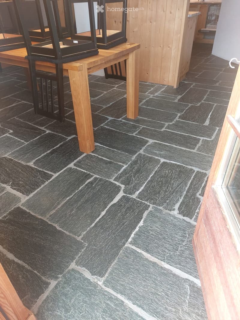 A room with a dark grey slate tiled floor, a wooden table with a black metal chair, and wooden walls.