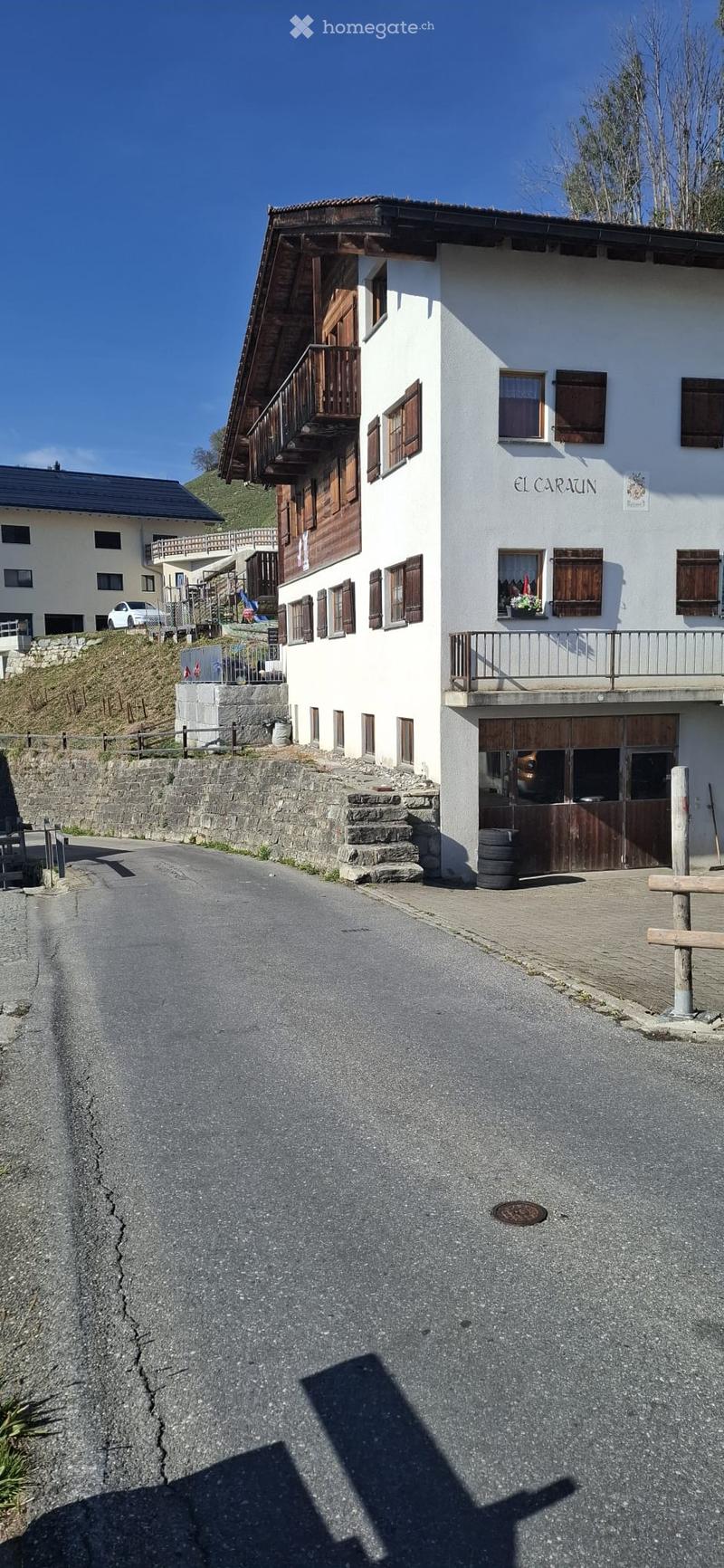 Two-story white building with wooden balconies and shutters, located on a narrow road with stone walls and a raised ground floor.