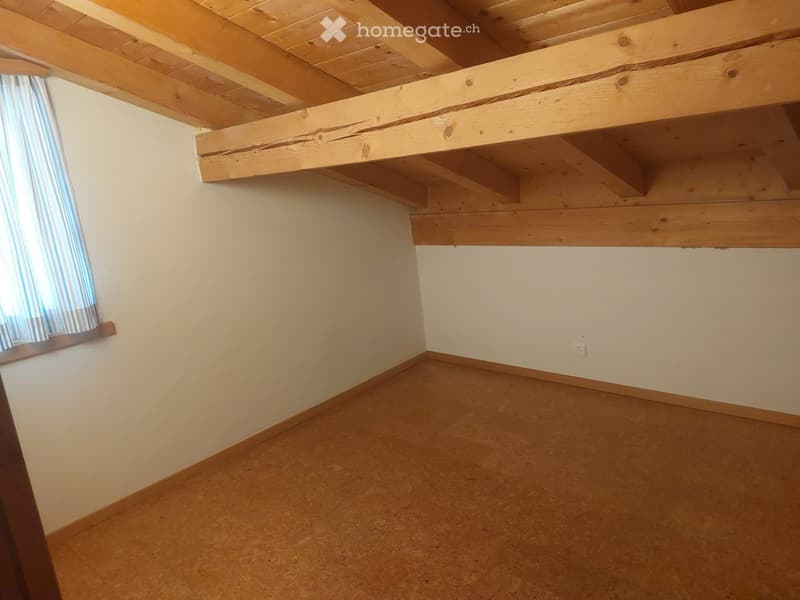 Empty room with exposed wooden beams on the ceiling, light-colored walls, and a window with striped curtains.