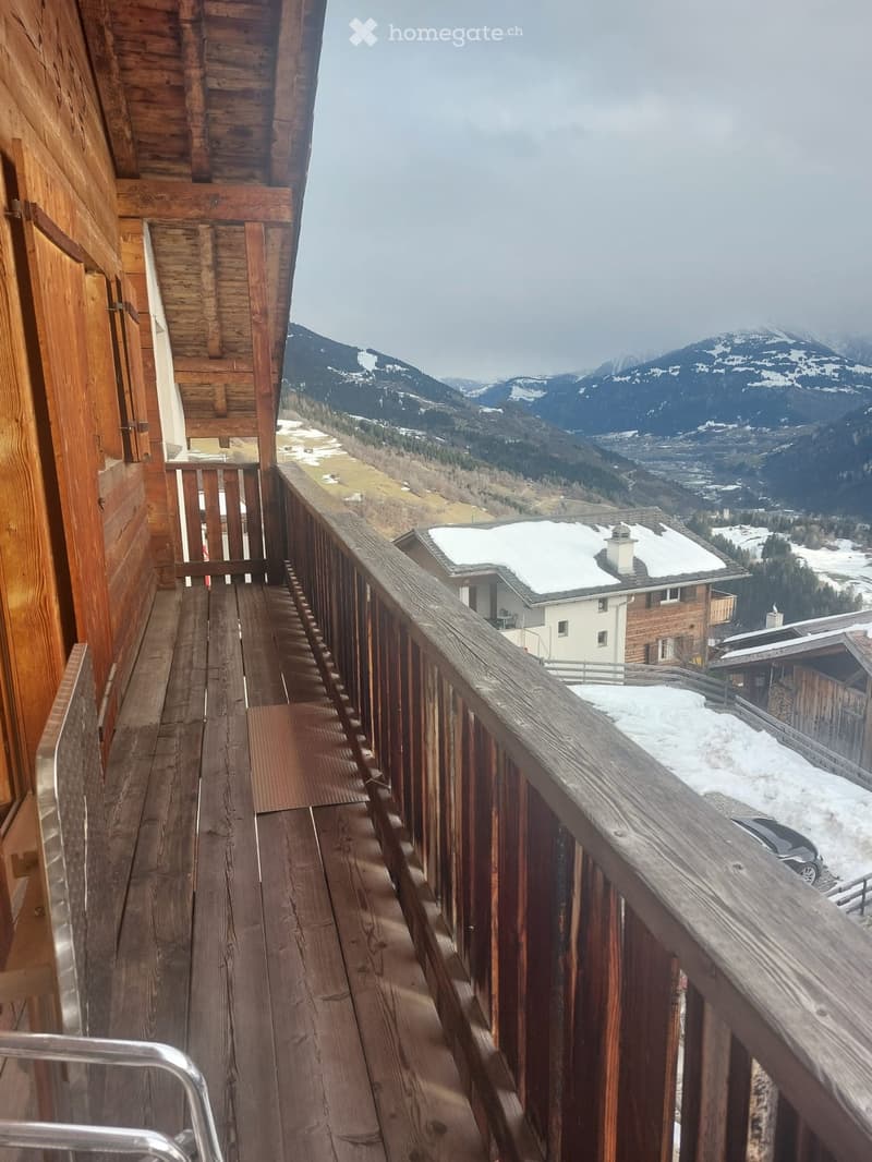 Wooden balcony with railing, overlooking snow-covered mountains and a village below.