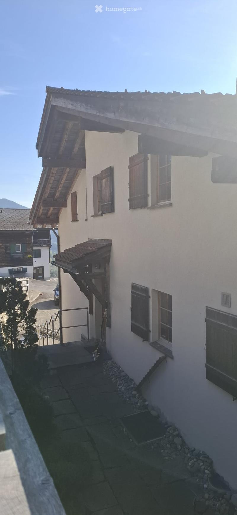 A two-story white building with wooden shutters and a wooden balcony, situated in a mountainous area with a clear blue sky and distant mountains.