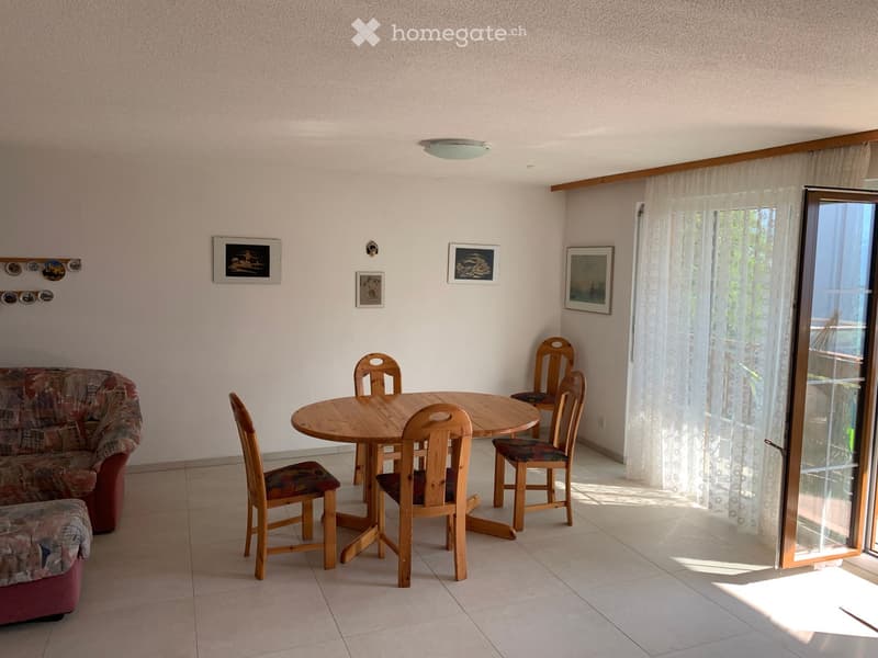 A cozy living and dining area with a red patterned sofa, ottoman, round wooden dining table, and six wooden chairs. The room features a large window with white curtains and several framed pictures on the wall.