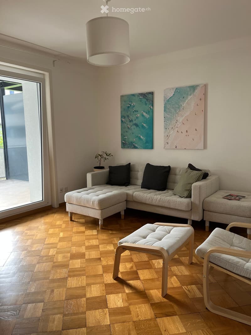 Modern living room with light grey sectional sofa, matching stool, and armchair. Features wooden flooring, pendant light, and two abstract paintings on the wall. Large window with sliding glass door provides natural light.