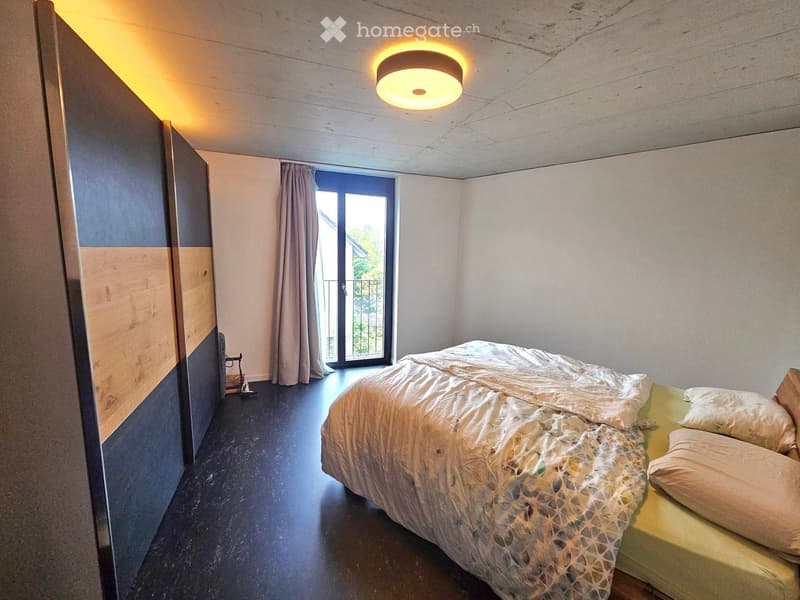 Modern bedroom with a bed, pillows, a wardrobe, a curtain, and a ceiling lamp. The room features a dark wooden floor and a large window with a view.