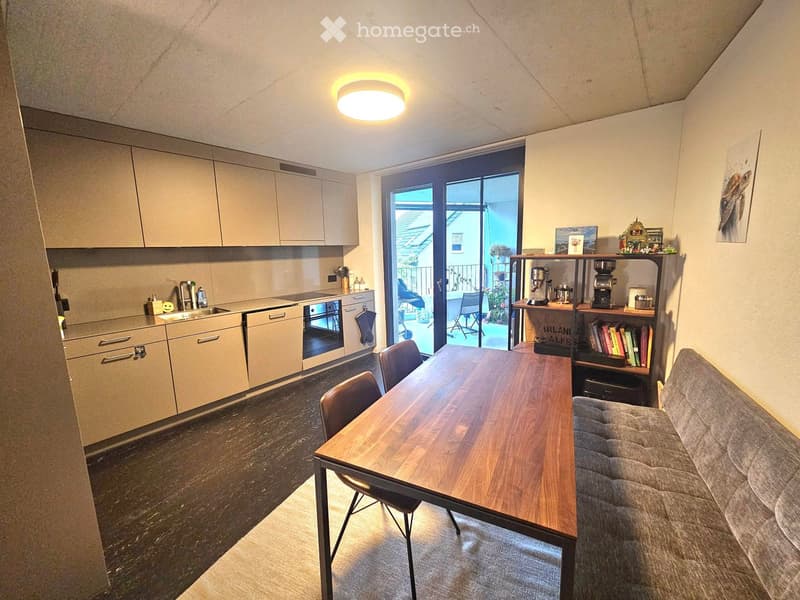 Modern open-plan living and dining area with a grey sofa, wooden dining table, chairs, kitchen area with cabinets, sink, oven, and dishwasher, and a bookshelf with coffee maker and books.