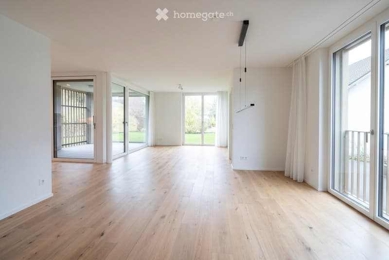 Bright, empty room with light wooden flooring, large glass sliding doors leading to a balcony, white walls, and modern lighting fixtures.