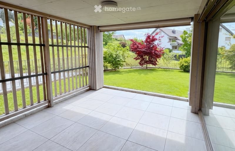 Spacious balcony with white tiled flooring, wooden railings, and large glass panels offering views of a well-maintained garden and surrounding houses.