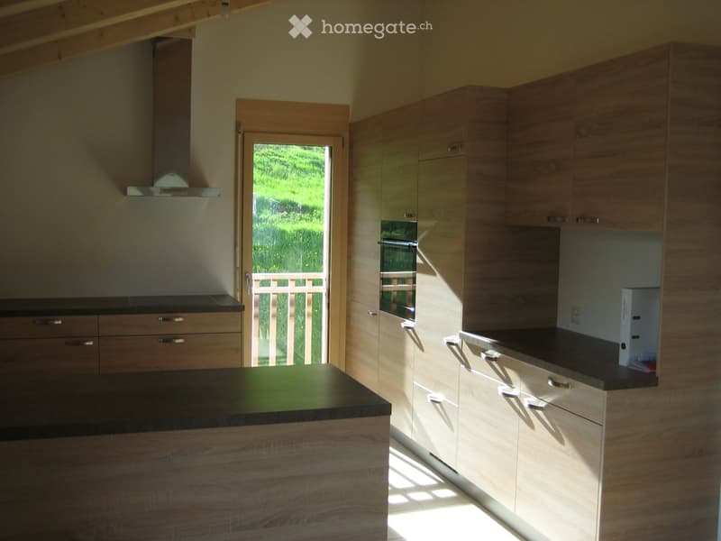 Modern kitchen with light wood cabinets, black countertops, and an island. Features an oven, range hood, and a door leading to a green outdoor area.
