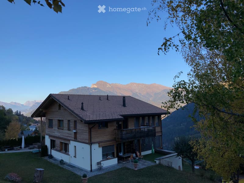 A two-story wooden house with a tiled roof, surrounded by a well-maintained garden and scenic mountain views.