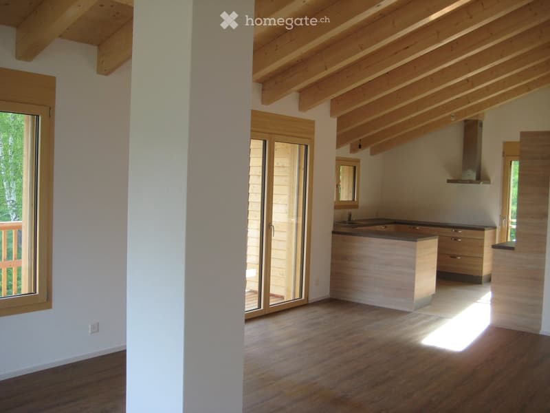 Open-plan space with wooden flooring, exposed wooden beams on the ceiling, large windows, and a kitchen area with cabinets and a sink.