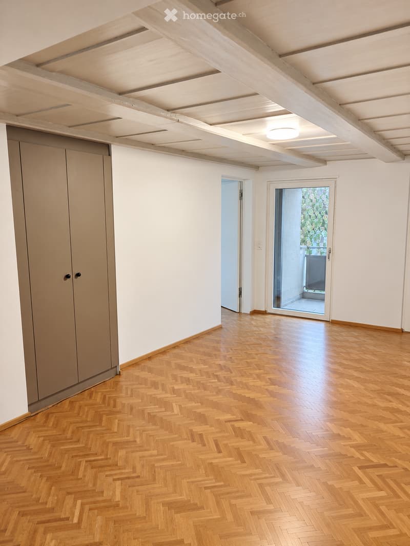 Empty room with wooden herringbone flooring, white walls, exposed wooden ceiling beams, and a sliding glass door leading to a balcony.