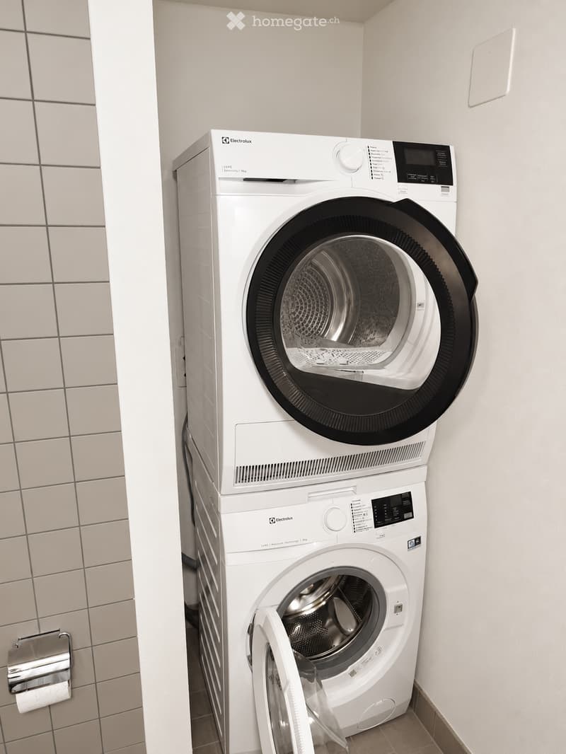 A compact bathroom with a stacked washing machine and dryer unit, white tiled walls, and a toilet paper holder visible on the left side.