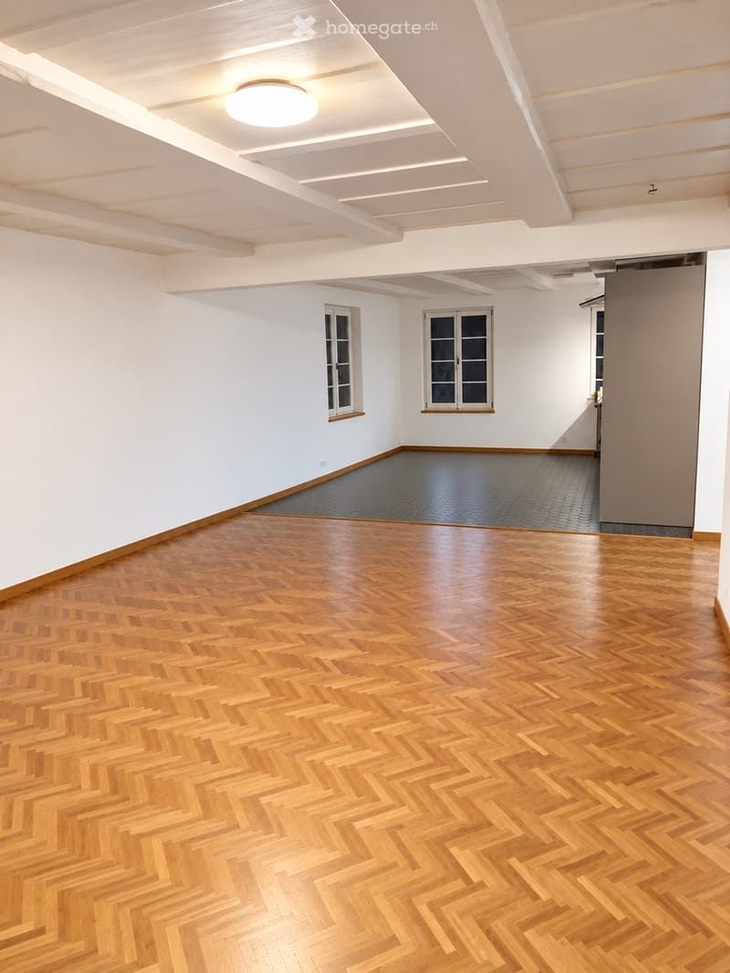 An empty room with polished wooden herringbone flooring, white walls, and large windows. The ceiling features recessed lighting and exposed beams.
