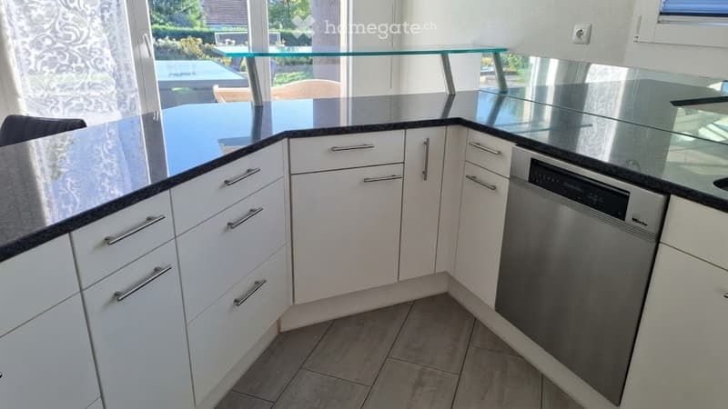 Modern kitchen with white cabinets, black countertops, and a built-in dishwasher. The space features a glass table and chairs visible through the open doorway.
