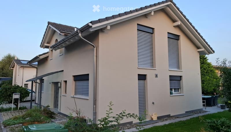 Two-story house with beige exterior, multiple windows with shutters, small garden area, and a covered entrance.