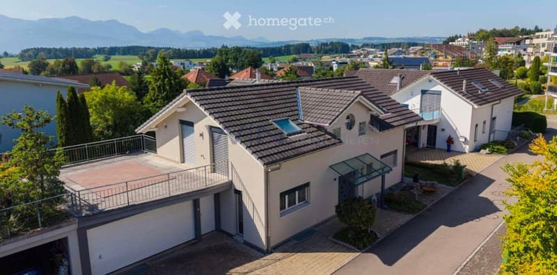 Aerial view of a modern two-story house with a tiled roof, garage, and surrounding greenery, set in a suburban neighborhood with distant mountains.