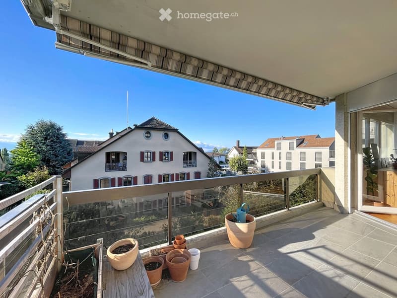 Balcony with tiled floor, glass railing, and several potted plants arranged on a small wooden table. A watering can is placed nearby.