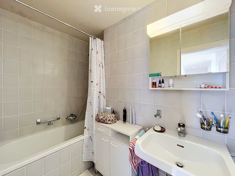 Modern bathroom with a bathtub, sink, mirror, and various toiletries. Features tiled walls and a shower curtain.