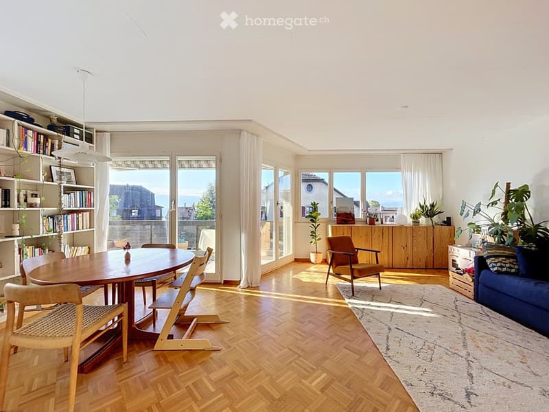 Bright, open living and dining area with wooden flooring, large windows, and a view. Features a blue sofa, wooden dining table with chairs, bookshelf, and potted plants.