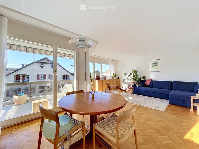 Bright living and dining area with wooden flooring, large windows, and a balcony. Features a blue sofa, round wooden dining table with chairs, pendant light, and decorative plants.