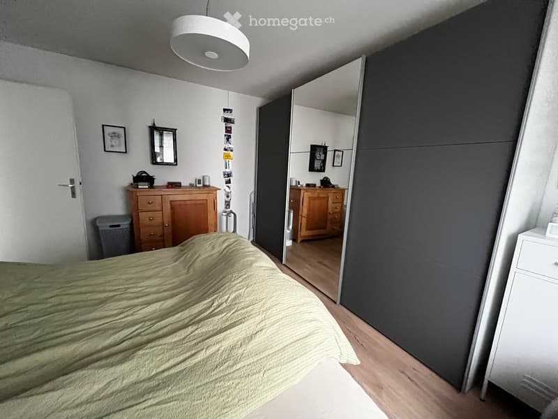 Bedroom with a large bed covered in a green duvet, a wooden wardrobe, a mirror, and a picture frame on the wall.