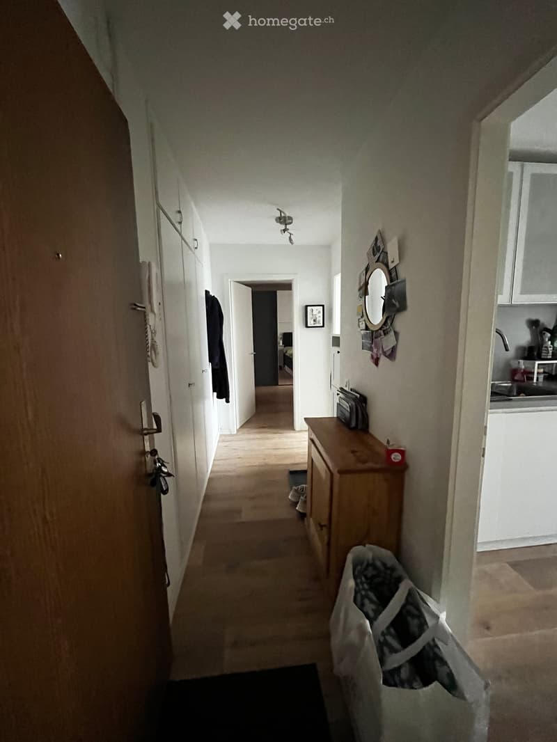 A narrow hallway with wooden flooring, white walls, and several doors leading to different rooms. A small wooden table with a mirror and some items is visible on the right side, and a coat is hanging on the left wall.