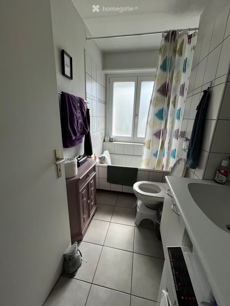 Bathroom with a toilet, bathtub, sink, and cabinet. Includes a shower curtain, towels, a framed picture, a wicker basket, and a small stool.